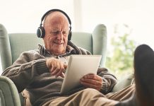 Happy elderly man relaxing on a chair at home and listening to music on digital tablet with headphones