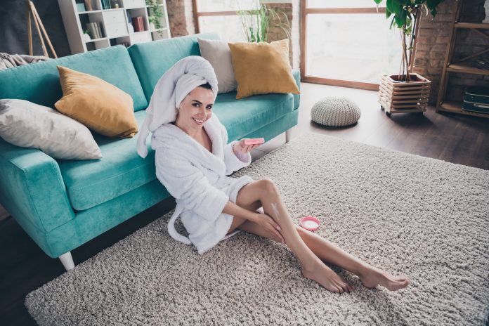 Portrait of attractive lady apply body cream lotion balm after legs waxing epilation sitting floor carpet spacious flat living room indoors